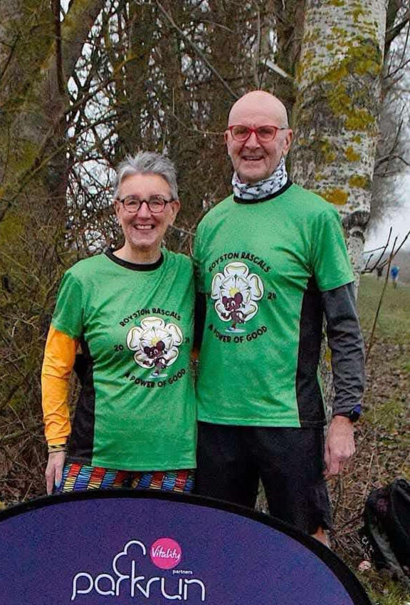 Man and woman standing side by side in club shirts, smiling at camera in front of ParkRun sign.